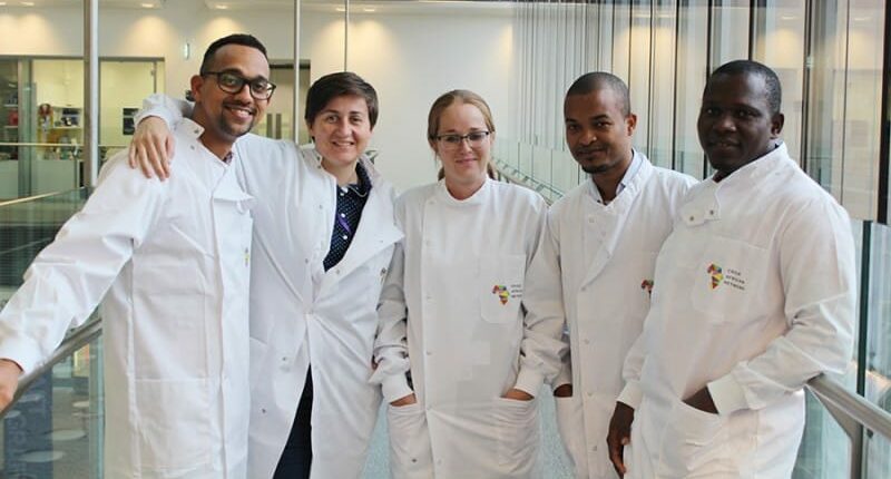 5 researchers in white lab coats stand together smiling with their arms around each other's shoulders.