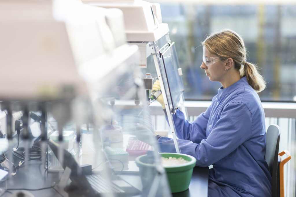 A female scientist is wearing PPE in a lab, carrying out an experiment