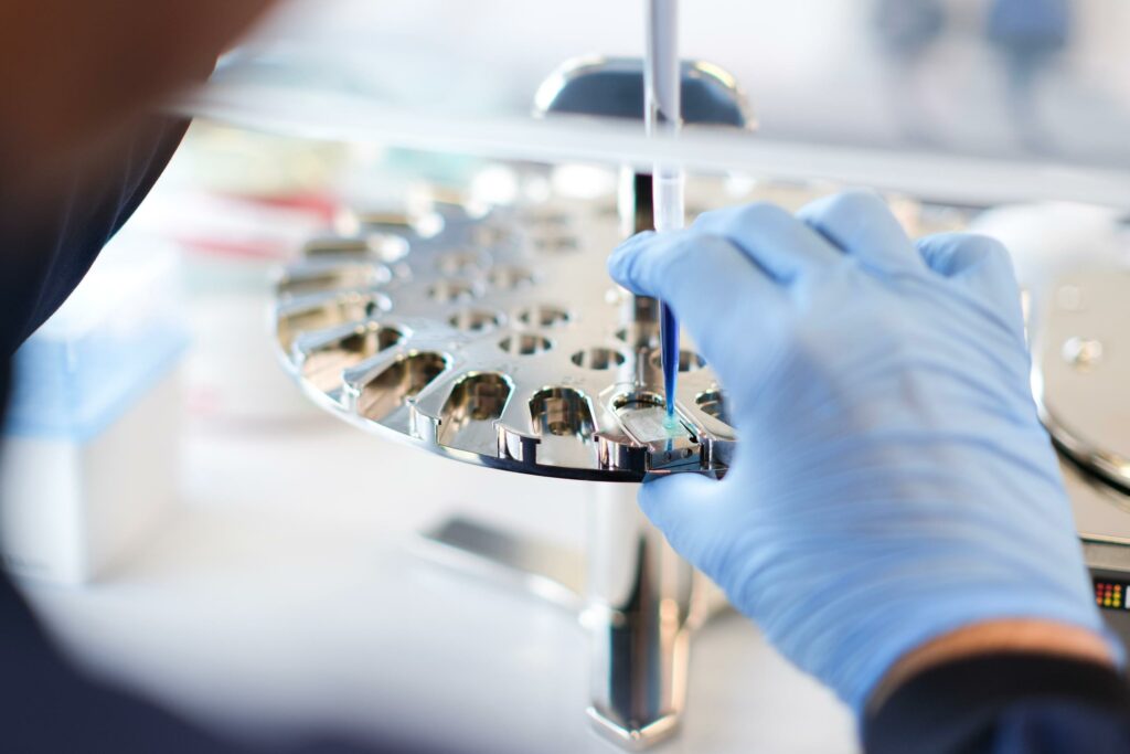 A gloved hand holds a pipette filled with blue liquid over a piece of lab equipment