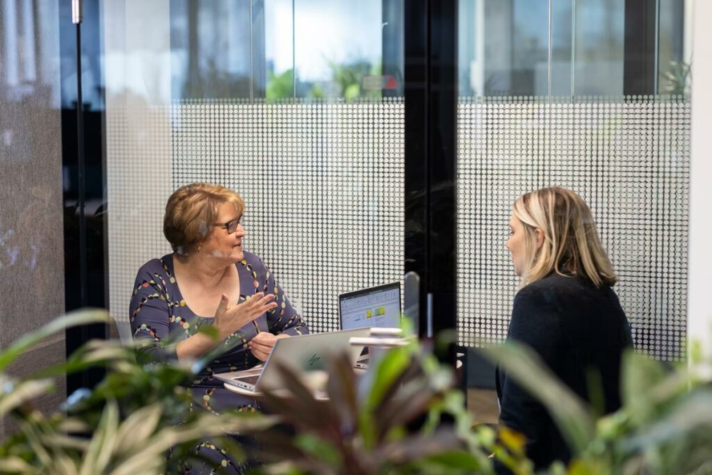 Two female LifeArc colleagues sit having a discussion in a glass office meeting pod