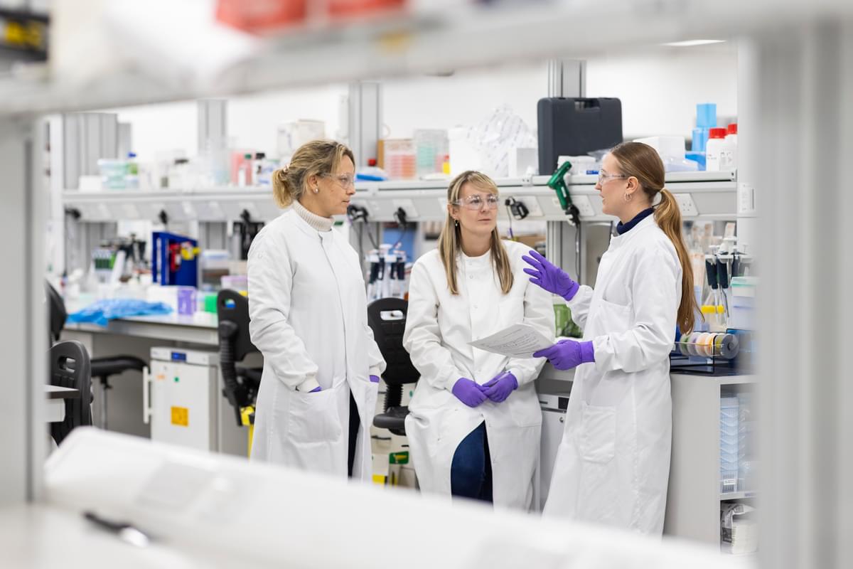 Three female scientists standing, having a discussion in the LifeArc lab.