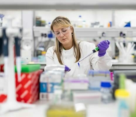 A female scientist is wearing PPE in a lab, holding a pipette and carrying out an experiment