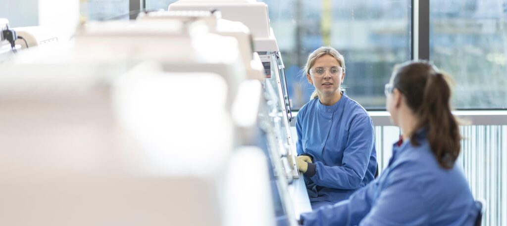 Two female scientists wearing PPE are sitting at a lab bench infront of lab equipment and are talking.