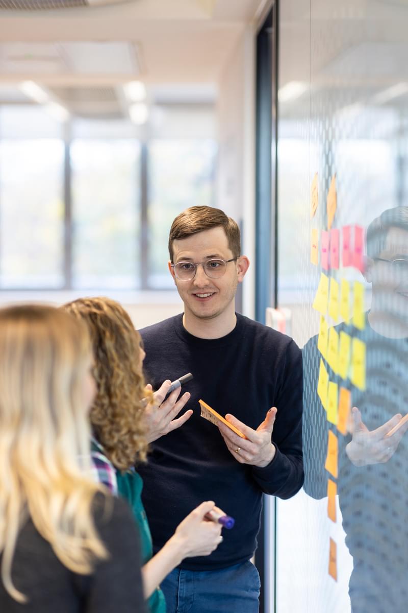 LifeArc Head of Policy and Public affairs talks to two colleagues as they brainstorm with post-it notes stuck to the wall