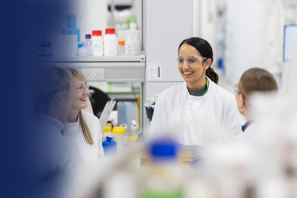 A group of four female LifeArc scientists stand smiling and talking to each other in a lab.