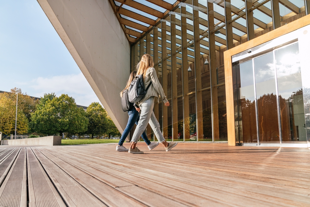 Two young females wearing casual clothing and backpacks walk towards the doors of a large modern building.