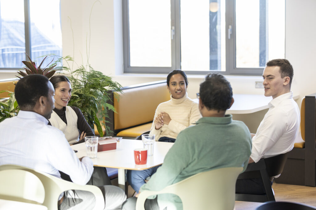 A group of LifeArc colleagues sit, in animated discussion, at a table in a communal office area,