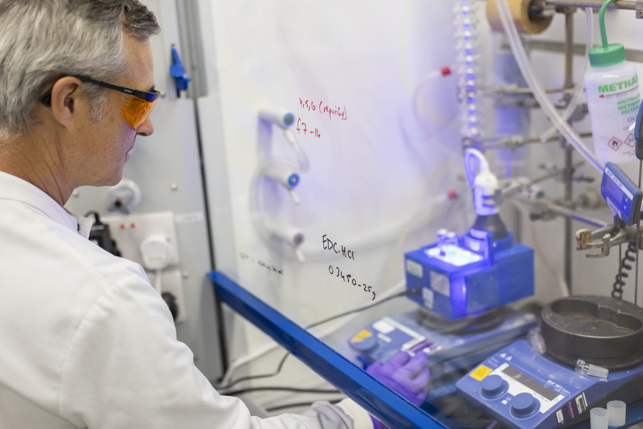A male scientist operates chemistry lab equipment behind a splash shield.