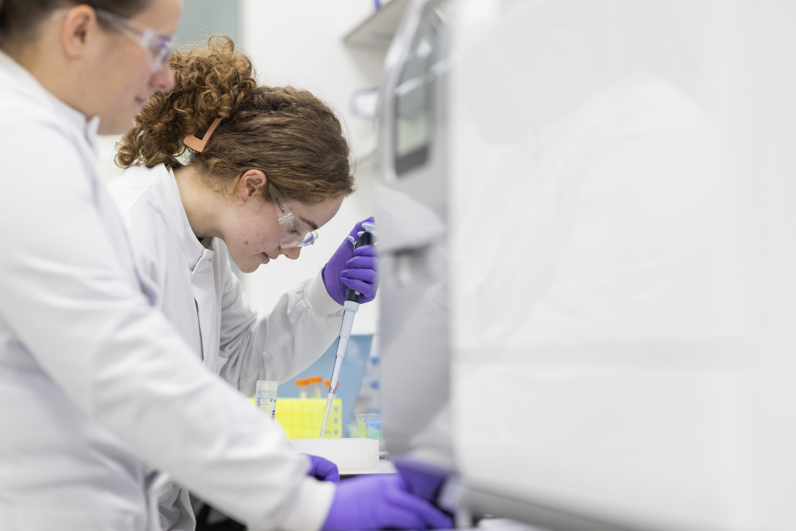 Two female scientists in lab coats and gloves work at a lab bench.