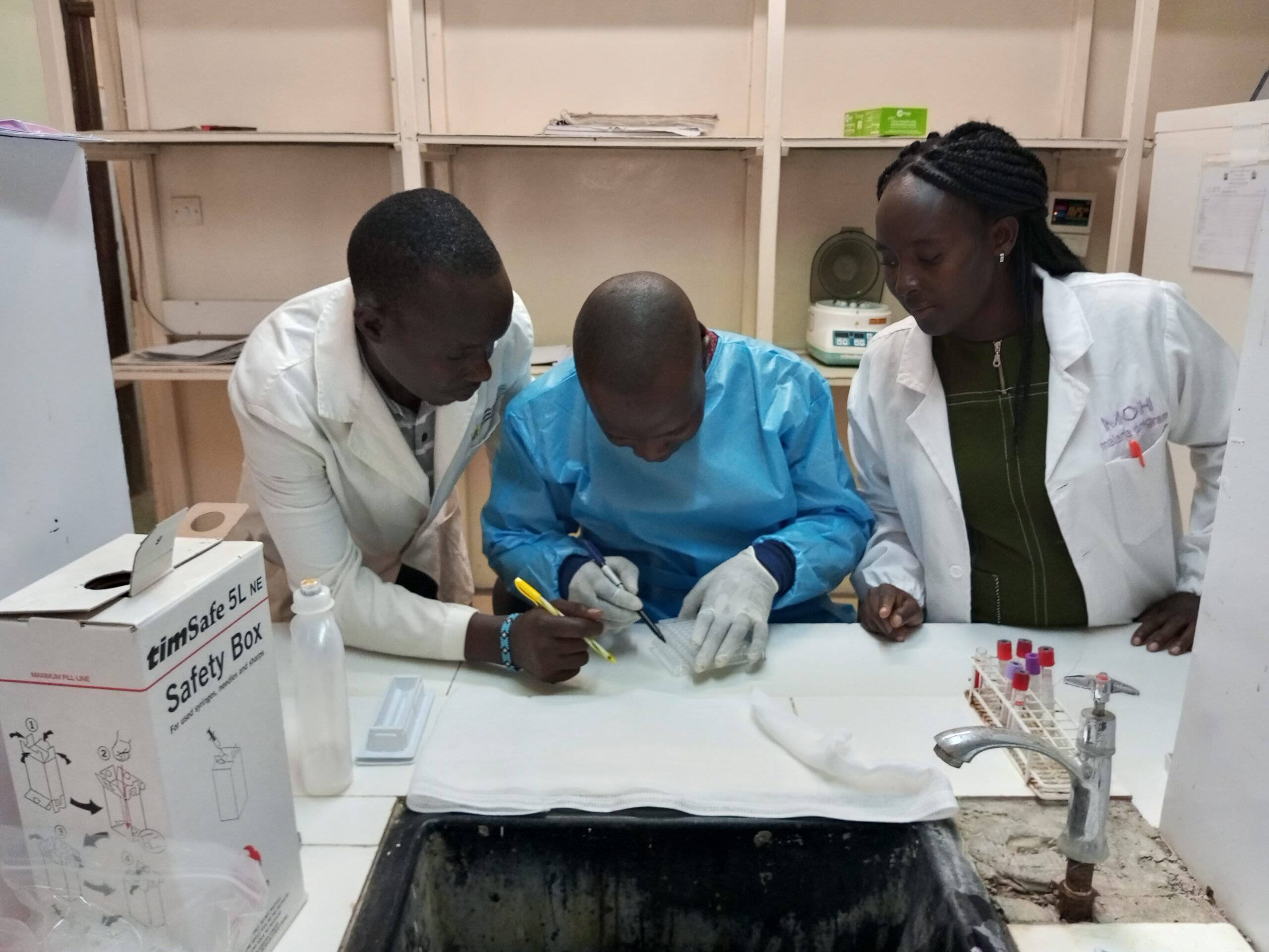 Three African scientists wearing lab coats and gloves looking at a scientific implement together