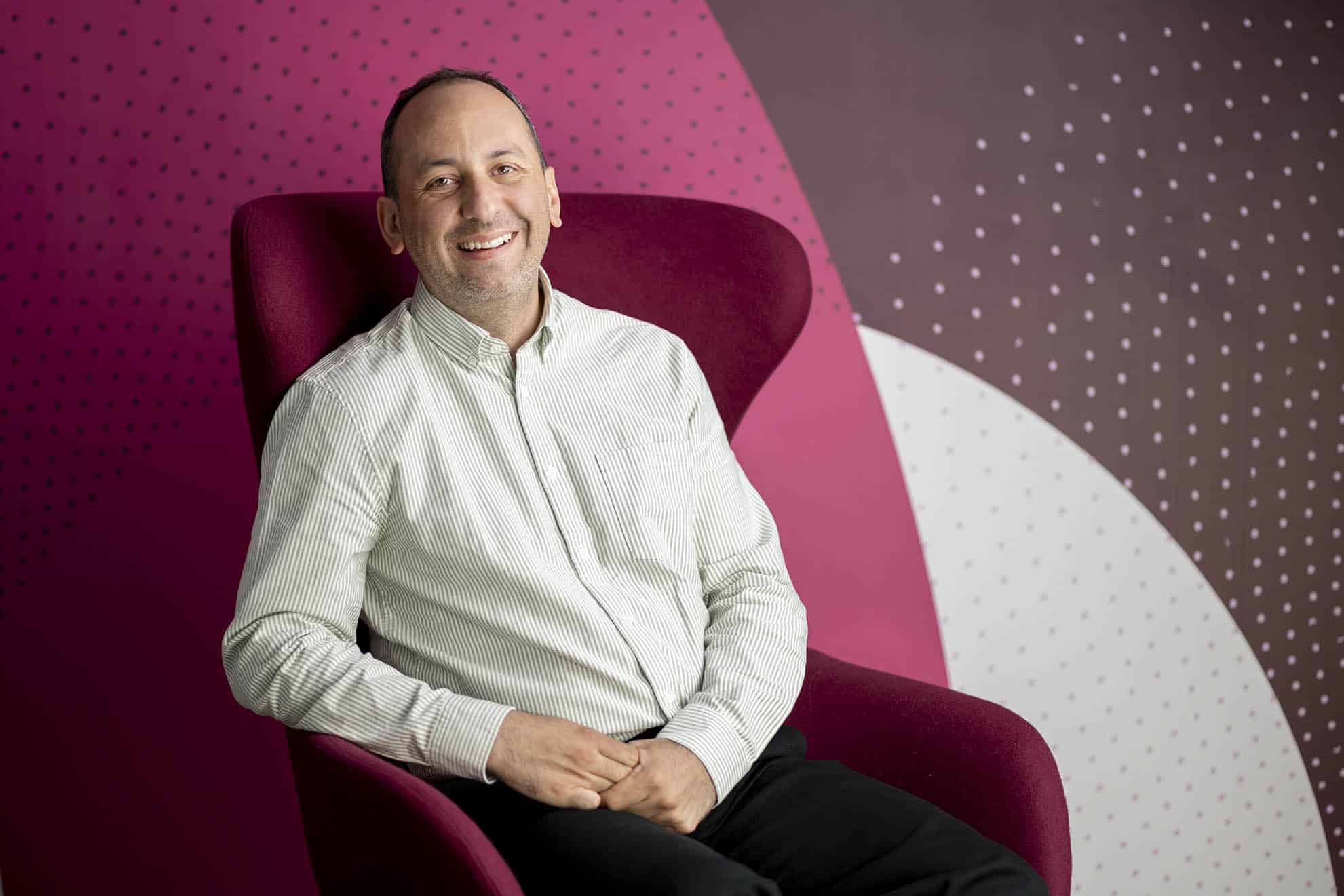 Photograph of Nessim Kichik sitting in a pink chair smiling at the camera