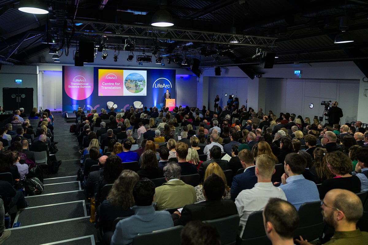 Crowded conference hall with a LifeArc branding on the stage and a LifeArc speaker