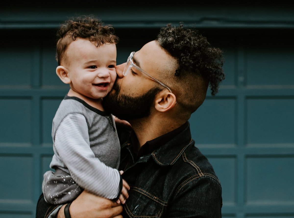 A young dad holding his toddler up and kissing him.