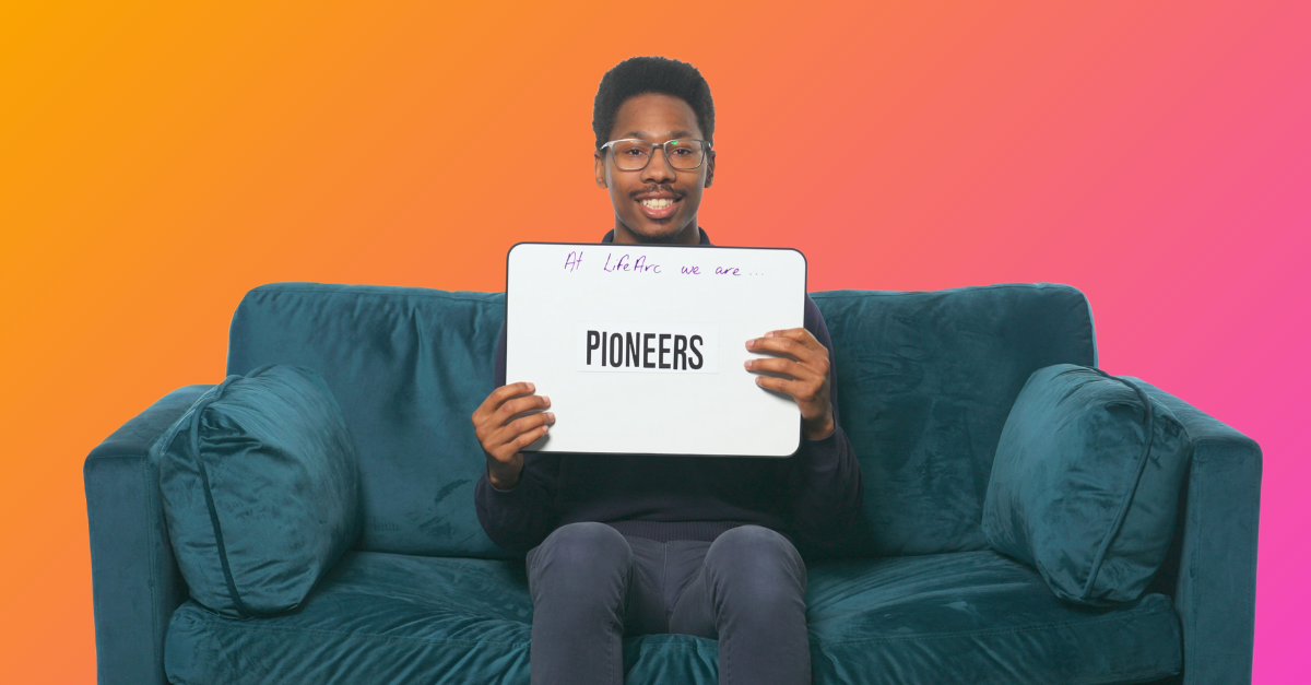 Asher, a LifeArc employee sits on a sofa holding up a sign saying 'pioneers'.