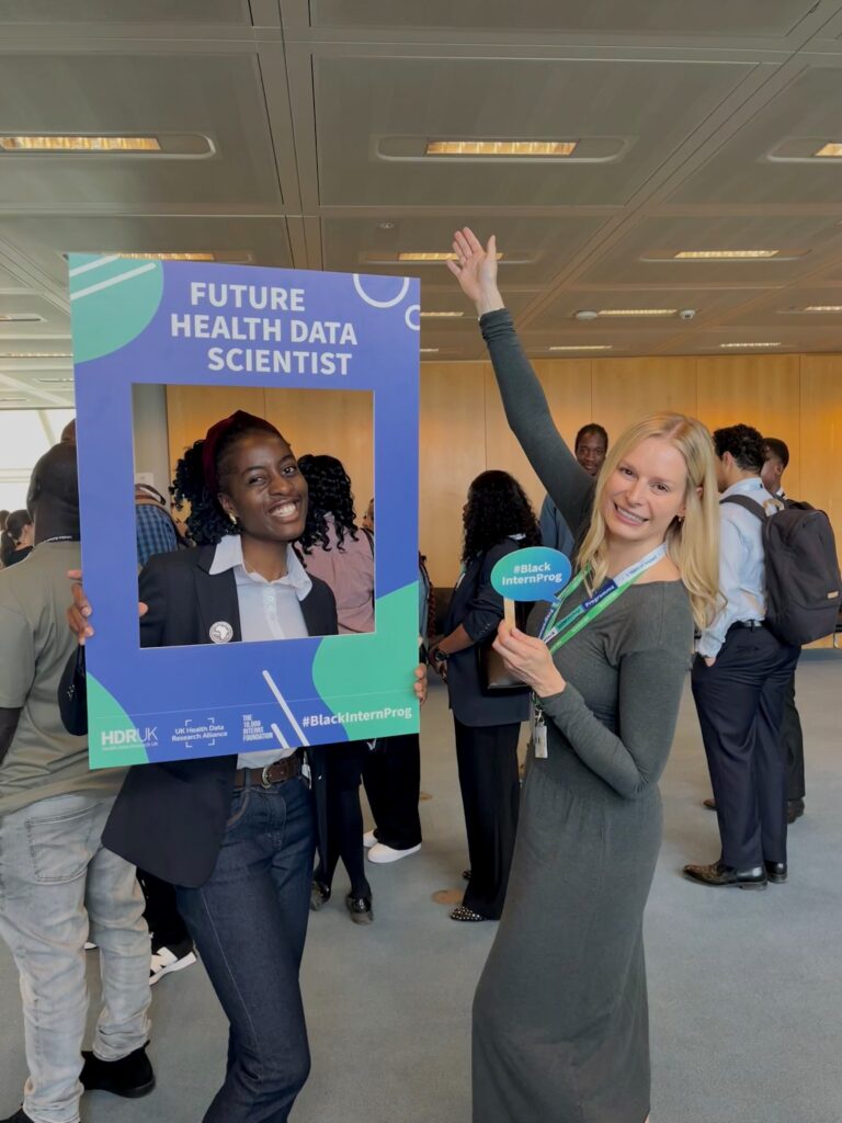 Varaidzo and LifeArc data scientist, Rebecca, are smiling and posing at an event. Rebecca holds a large blue frame with the text 'FUTURE HEALTH DATA SCIENTIST' and '#BlackInternProg'. Varaidzo stands beside them, raising one arm and wearing a lanyard with a '#BlackInternProg' badge.