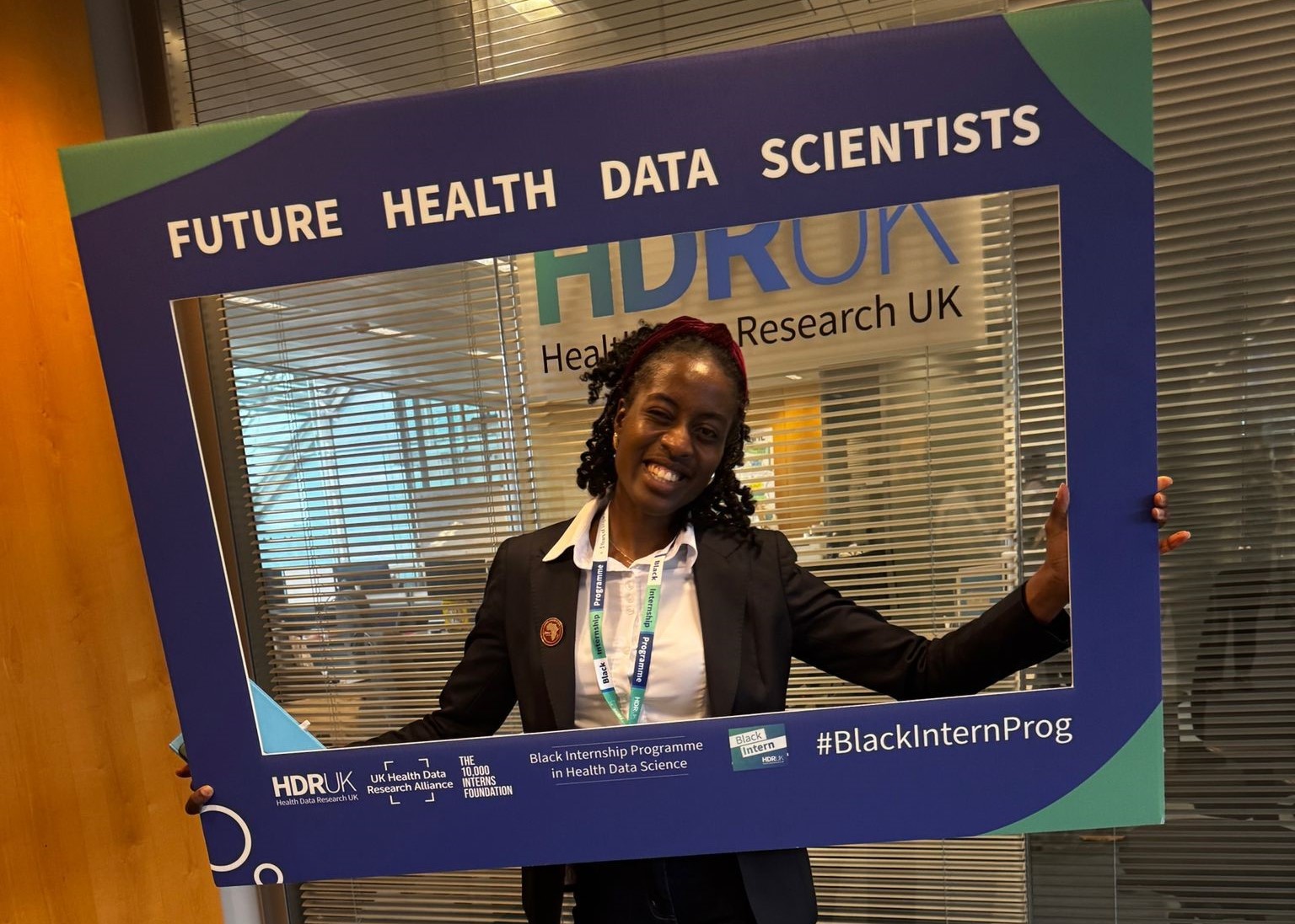 Varaidzo smiling and holding a large blue frame that reads 'FUTURE HEALTH DATA SCIENTISTS'. The frame features logos for HDR UK, Health Data Research UK, and the Black Internship Programme in Health Data Science.
