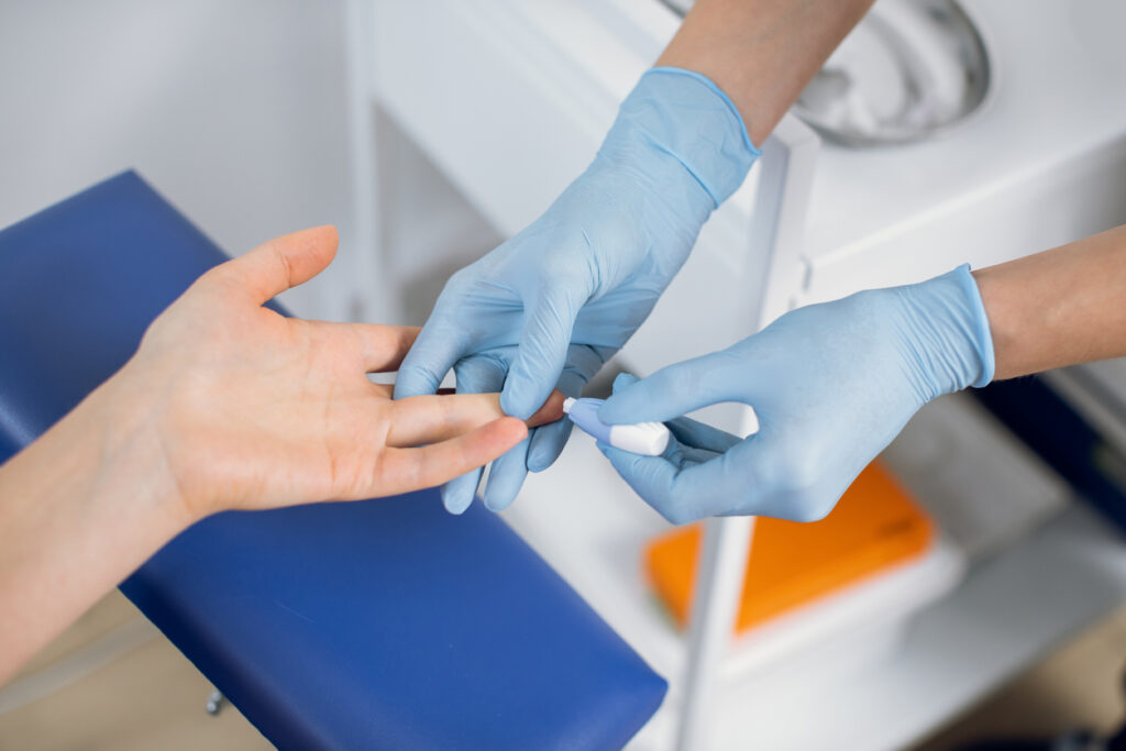 Close up of nurse lab technician in blue sterile gloves, using painless scarifier to prick the finger of unrecognizable patient, sitting on chair. Blood analysis and group determination