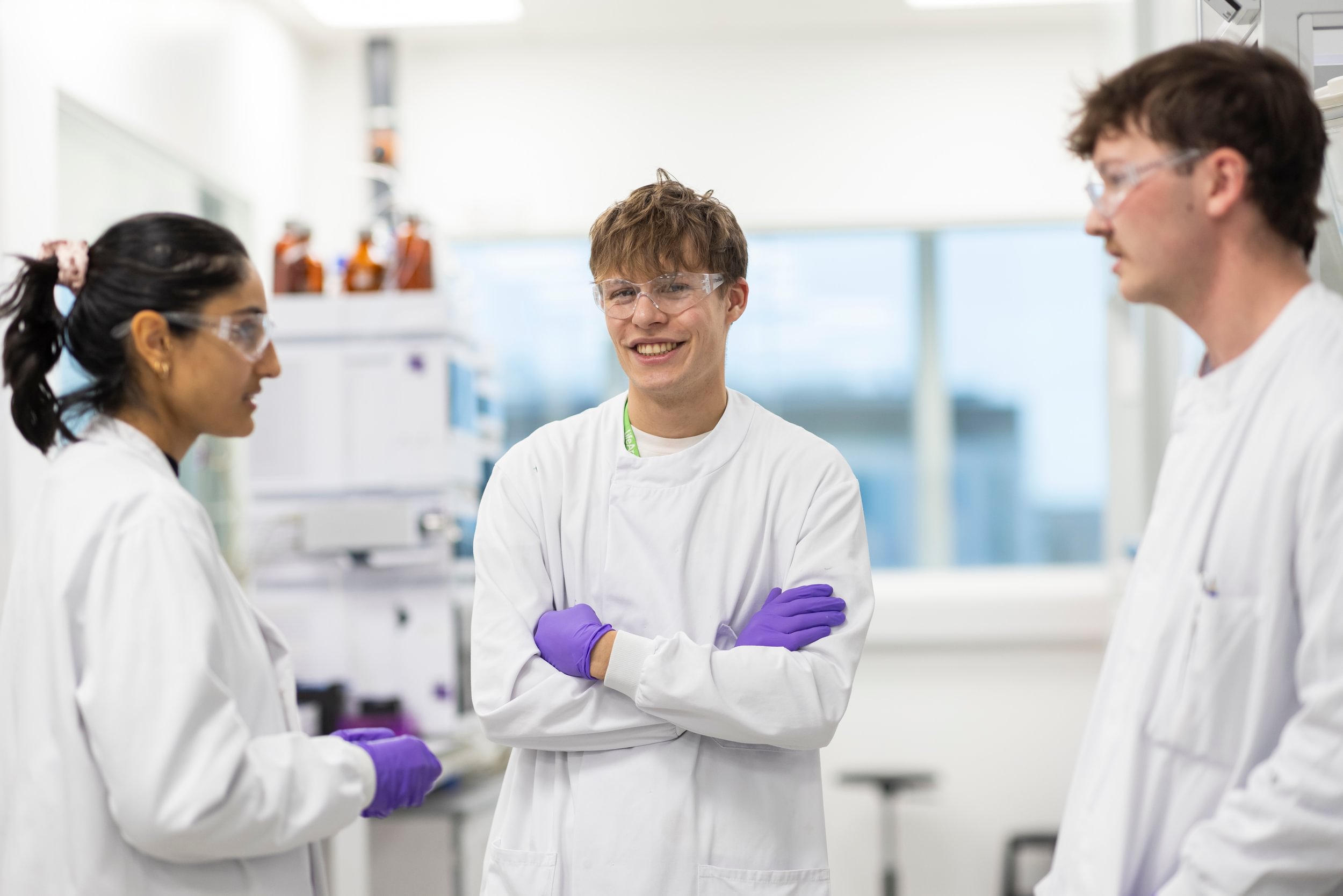 Three LifeArc scientists, two male and one female, wearing lab coats and gloves, stand in a lab talking.