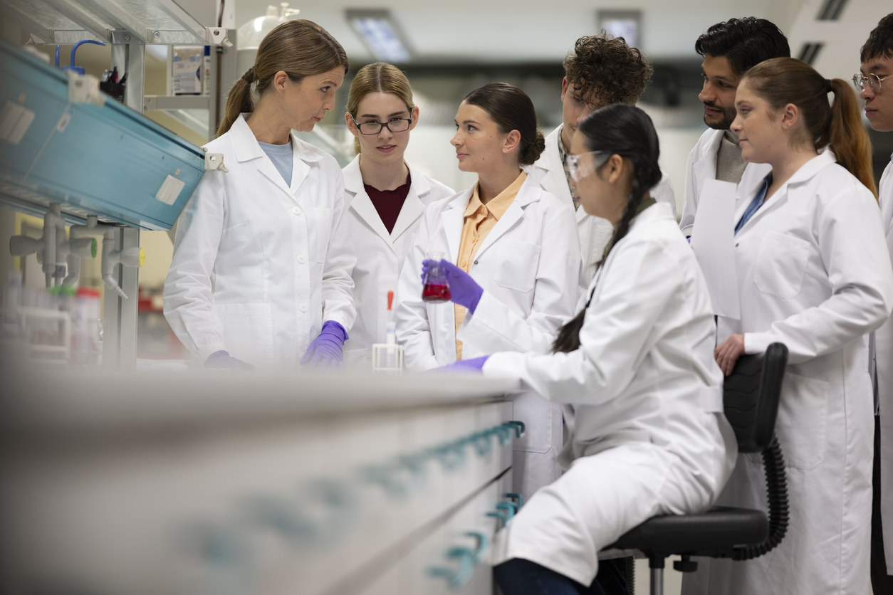 Female professor supervising a diverse group of students in a laboratory setting. Students observing experiment and holding beaker with red liquid. Learning and scientific productivity taking place in lab.